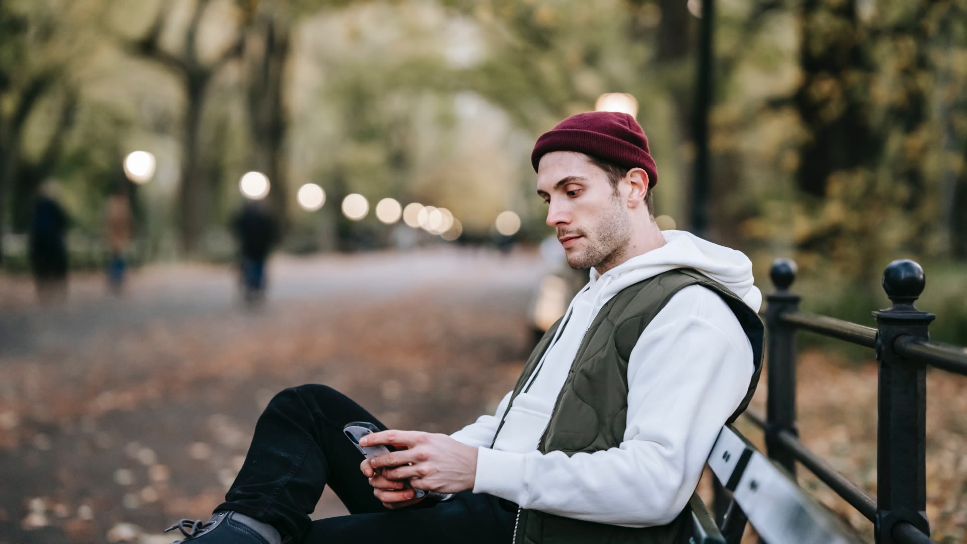 A young man in a white hoodie, olive vest, and maroon beanie looks at his phone while sitting on a park bench.