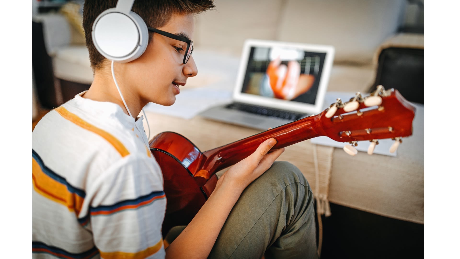A young teenager in a striped shirt plays guitar while listening to headphones.
